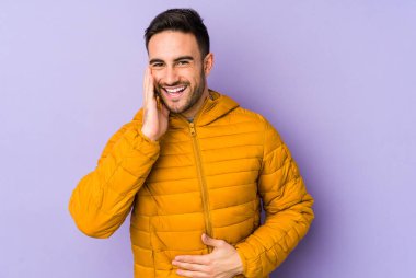 Mor arka planda izole edilmiş genç beyaz adam mutlu bir şekilde gülüyor ve ellerini karnının üstünde tutarak eğleniyor..