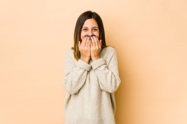 Bej arka planda izole edilmiş genç bir kadın bir şeye gülüyor, elleriyle ağzını kapatıyor..