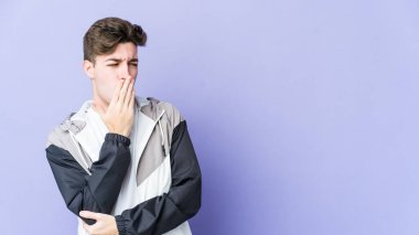 Mor arka planda izole edilmiş genç beyaz adam esniyor. Ağzı eliyle kapatan yorgun bir hareket sergiliyor..
