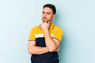 Kafkasyalı genç adam mavi arka planda izole edilmiş şüpheli, belirsiz, seni muayene ediyor..