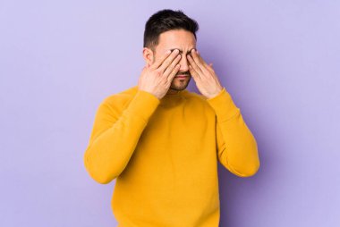 Mor arka planda izole edilmiş genç beyaz adam elleriyle gözlerini kapatmaktan korkuyor..