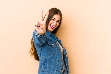 Beyaz tenli genç bir kadın bej arka planda izole edilmiş neşeli ve kaygısız parmaklarıyla barış sembolü gösteriyor..