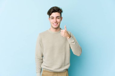 Mavi arka planda izole edilmiş genç beyaz adam gülümsüyor ve başparmağını kaldırıyor.