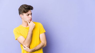 Mor arka planda izole edilmiş genç beyaz adam şüpheli ve şüpheci bir ifadeyle yana bakıyor..