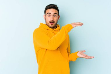 Mavi arka planda izole genç beyaz adam şok ve hayret içinde elleri arasında bir kopya boşluk tutuyor.