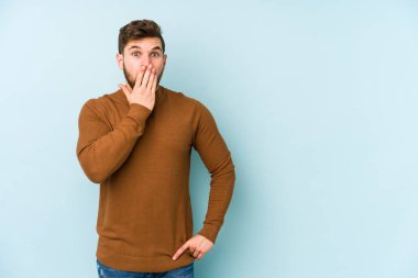 Mavi arka planda izole edilmiş genç beyaz adam şok olmuş, ağzı ellerle kaplanmış, yeni bir şey keşfetmeye hevesli..