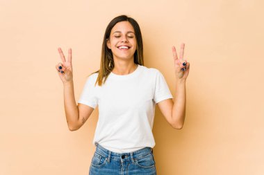 Bej arka planda izole edilmiş genç bir kadın zafer işareti gösteriyor ve gülümsüyor..