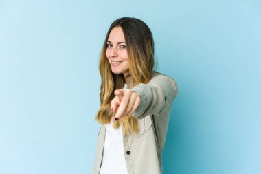 Mavi arka planda izole edilmiş genç beyaz kadın parmaklarıyla ön tarafı işaret ediyor..