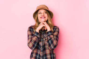 Şapka takan genç beyaz kadın ellerini çenesinin altında tutuyor ve mutlu bir şekilde yan tarafa bakıyor..