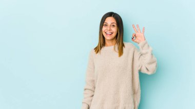 Mavi arka planda izole edilmiş genç bir kadın bir göz kırpıyor ve elleriyle iyi bir jest yapıyor..