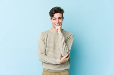 Mavi arka planda izole edilmiş genç beyaz adam gülümsüyor ve kendinden emin, elleriyle çenesine dokunuyor..