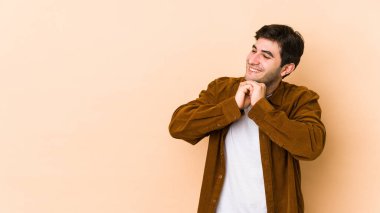 Bej arka planda izole genç adam ellerini çenesinin altında tutuyor, mutlu bir şekilde yana bakıyor..