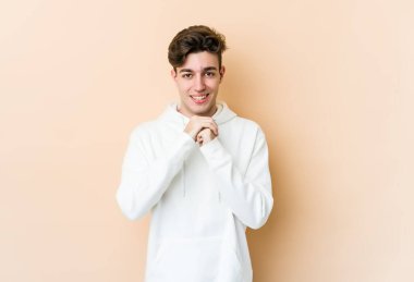 Beyaz tenli genç adam bej arka planda izole edilmiş ellerini çenesinin altında tutuyor, mutlu bir şekilde yana bakıyor..