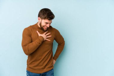 Kafkasyalı genç adam mavi arka planda izole olmuş gülüyor, elini yüreğine koyuyor, mutluluk kavramını koruyor..