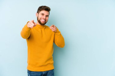 Mavi arka planda izole edilmiş genç beyaz adam parmaklarıyla ön tarafı işaret ediyor..