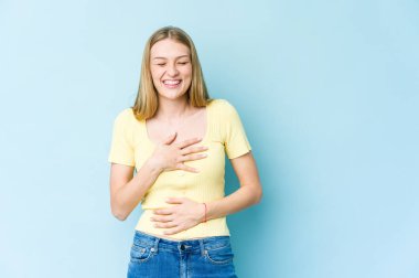 Mavi arka planda izole edilmiş genç sarışın kadın mutlu bir şekilde gülüyor ve ellerini karnının üstünde tutarak eğleniyor..