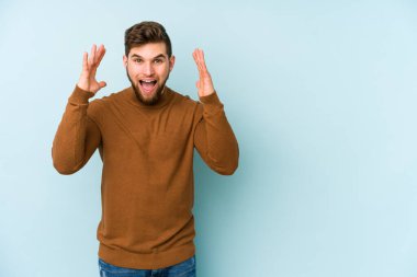 Mavi arka planda izole edilmiş genç beyaz adam hoş bir sürpriz alıyor, heyecanlı ve el kaldırıyor..