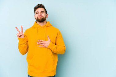 Kafkasyalı genç adam mavi arka planda izole edilmiş yemin ediyor, elini göğsüne koyuyor..