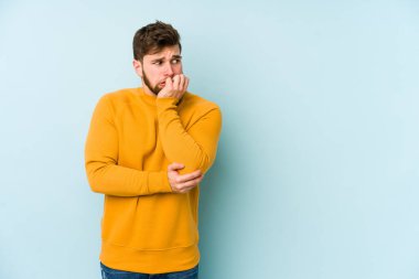 Mavi arka planda izole edilmiş, tırnak yiyen, gergin ve endişeli genç bir beyaz adam..