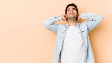 Genç adam bej arka planda izole edilmiş. Kendine güveniyor, elleri başının arkasında..