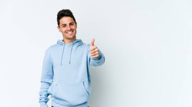 Beyaz arka planda izole edilmiş genç beyaz adam gülümsüyor ve başparmağını kaldırıyor.