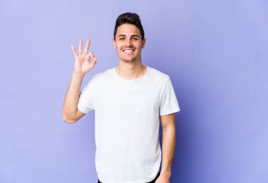 Mor arka planda izole edilmiş genç beyaz bir adam bir göz kırptı ve elleriyle iyi bir jest yaptı..