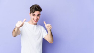 Mor arka planda izole edilmiş genç beyaz adam iki parmağını da kaldırıyor, gülümsüyor ve kendinden emin..