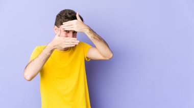 Mor arka planda izole edilmiş genç beyaz adam kameraya parmaklarıyla göz kırpıyor, yüzünü kapatmaktan utanıyor..