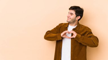 Bej arka planda izole edilmiş genç adam gülümsüyor ve elleriyle kalp şeklini gösteriyor..
