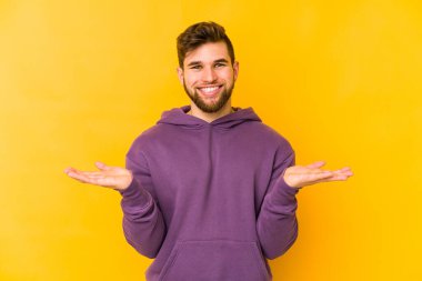 Sarı arka planda izole genç beyaz adam kollarıyla ölçek yapar, mutlu ve kendinden emin hisseder..