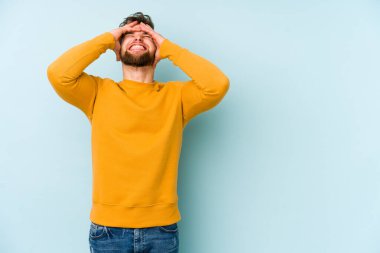 Mavi arka planda izole edilmiş genç beyaz adam ellerini başının üstünde tutarak neşe içinde gülüyor. Mutluluk kavramı.