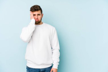 Kafkasyalı genç adam mavi arka planda izole edilmiş. Yorgun ve uykulu. Elini başının üstünde tutuyor..
