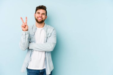 Mavi arka planda izole edilmiş genç beyaz adam iki numarayı parmaklarıyla gösteriyor..