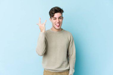 Mavi arka planda izole edilmiş genç beyaz adam parmaklarıyla kaya hareketi gösteriyor.