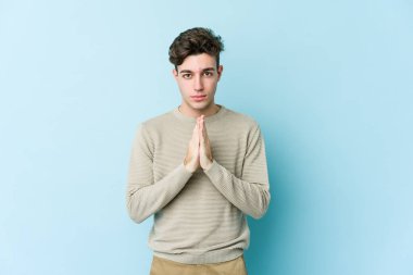 Kafkasyalı genç adam mavi arka planda izole edilmiş dua ediyor, bağlılık gösteriyor, dindar biri ilahi ilham arıyor..