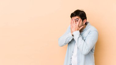 Bej arka planda izole edilmiş genç adam parmaklarının arasından korkmuş ve gergin bir şekilde göz kırp..