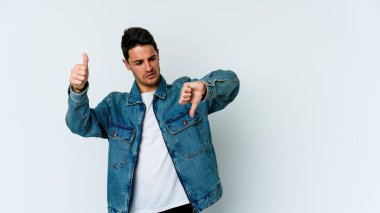 Beyaz arka planda izole edilmiş genç beyaz bir adam baş parmaklarını aşağı ve yukarı kaldırıyor, zor seçim konsepti