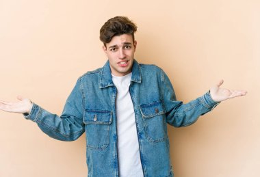 Kafkasyalı genç adam bej arka planda izole edilmiş. Kafası karışmış ve omuz silkiyor. Kopya alanını tutmak için..