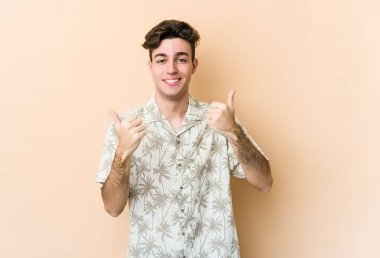 Beyaz tenli genç adam bej arka planda izole edilmiş. Baş parmağını kaldırıyor, gülümsüyor ve kendinden emin..