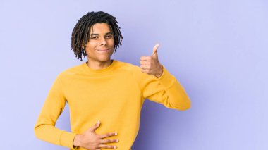 Rasta saç stili giyen genç siyahi adam karnına dokunuyor, nazikçe gülümsüyor, yemek yiyor ve tatmin oluyor..