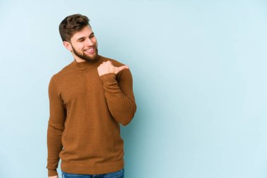 Kafkasyalı genç adam mavi arka plan noktalarında izole edilmiş başparmak uzakta, gülüyor ve kaygısız.