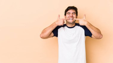 Bej arka planda izole edilmiş genç adam gülümsüyor, parmaklarını ağzına doğrultuyor..