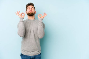 Mavi arka planda izole edilmiş genç beyaz adam yorucu bir günün ardından rahatlıyor. Yoga yapıyor..