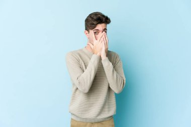 Kafkasyalı genç adam mavi arka planda izole edilmiş. Korkmuş ve gergin parmaklarıyla göz kırp..