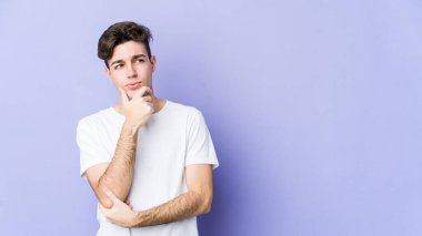 Kafkasyalı genç bir adam mor arka planda izole edilmiş düşünüyor, bakıyor, yansıtıyor, düşünüyor, bir fantezi kuruyor..