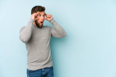 Mavi arka planda izole edilmiş genç beyaz adam bir başarı fırsatı bulmak için gözlerini açık tutuyor..