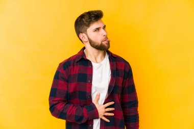 Sarı arka planda izole edilmiş genç beyaz adam karnına dokunur, nazikçe gülümser, yemek yer ve tatmin olur..