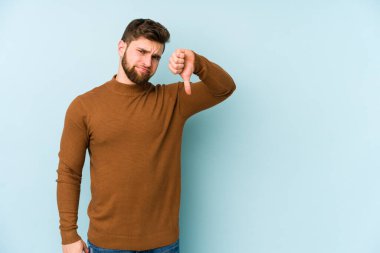 Mavi arka planda izole genç beyaz adam baş parmağını aşağı gösteriyor, hayal kırıklığı konsepti.