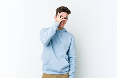 Beyaz arka planda izole edilmiş genç beyaz bir adam kameraya parmaklarıyla göz kırpıyor, yüzünü kapatmaktan utanıyor..