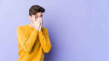 Mor arka planda izole edilmiş genç beyaz bir adam parmaklarının arasından korkmuş ve gergin bir şekilde göz kırpıyor..